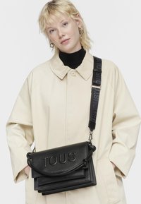 Borsa a tracolla in pelle nera con logo "TOUS" in rilievo, dotata di patta ribaltabile, manico annodato e una tracolla con marchio stampato in nero.