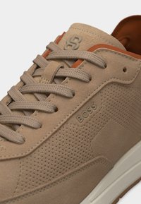 Baskets en daim beige avec perforations sur le côté, lacets beiges, bordure marron clair, logo "BOSS" embossé sur le côté et détails de couture superposée.