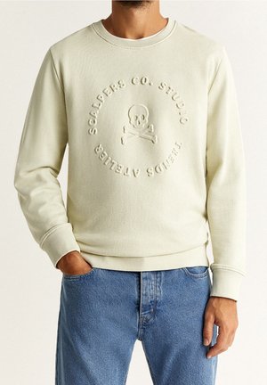 Man wearing a cream sweatshirt with embossed skull and crossbones and circular text, paired with blue jeans, hand in pocket.