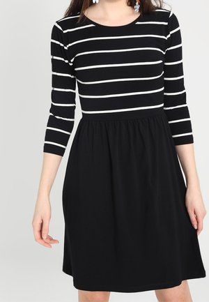 Woman wearing a black dress with white horizontal stripes on the top and three-quarter sleeves, plain black skirt, standing with arms relaxed.