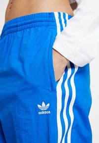 Siniset urheilushortsit, jotka on valmistettu kevyestä kankaasta. Shortsien sivuilla on valkoinen Adidas-logo ja kolme pystysuoraa valkoista raitaa.