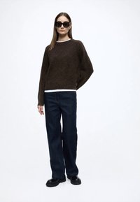 Hnedý sveter s textúrou, navrstvený na bielej košeli, v kombinácii s tmavomodrými džínsami širokým strihom a čiernymi robustnými topánkami, modelka má na sebe slnečné okuliare.