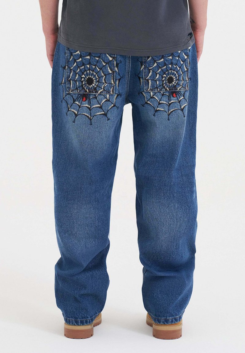 Osoba w niebieskich dżinsach z haftowanymi pajęczynami i małymi czerwonymi pająkami na tylnych kieszeniach, stojąca na białym tle.