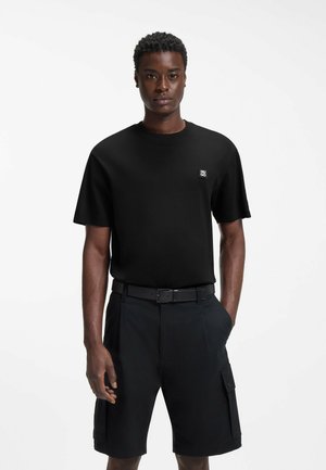 Junger Mann trägt schwarzes kurzärmeliges Shirt mit kleinem Logo und schwarze Cargo-Shorts mit Gürtel, steht vor einfachem weißem Hintergrund.