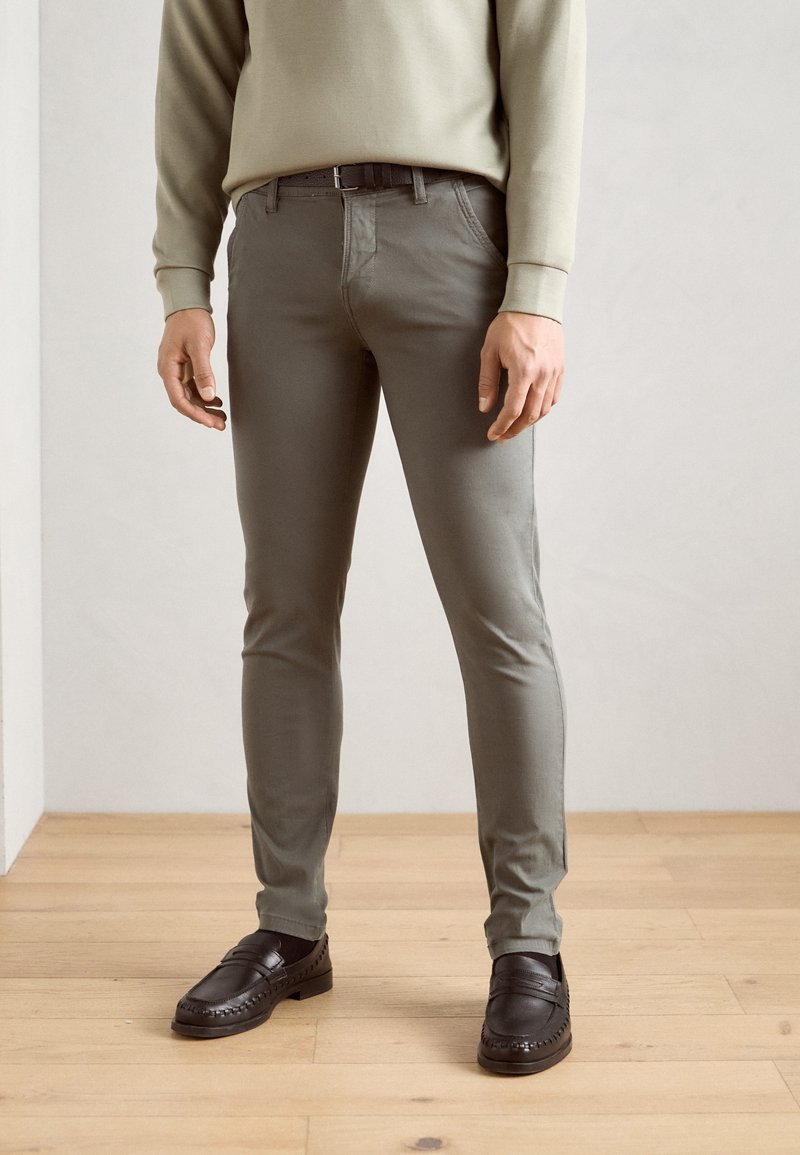 Homme portant un pantalon slim-fit vert olive, un haut beige à manches longues, une ceinture noire, des chaussettes noires et des mocassins noirs, debout sur un sol en bois.