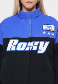 Czarna i niebieska kurtka z dużym, białym napisem "Roxy", teksturowanym materiałem, wysokim kołnierzem oraz naszywką z wyszytymi trzema gwiazdami.