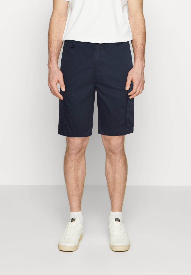 Homme portant un short cargo bleu marine, des chaussettes blanches, des baskets blanches et un t-shirt blanc sur un fond clair uni.