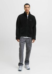 Chaqueta de forro polar negra con cremallera frontal, mangas largas y tejido texturizado; combinada con pantalones cargo gris claro y zapatillas deportivas plateadas.