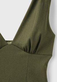 Gros plan d'une robe sans manches texturée vert olive avec un détail froncé au niveau de la poitrine et de larges bretelles.