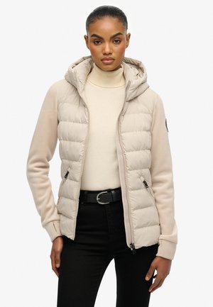 Gewatteerde lichtbeige jas met capuchon en gladde mouwen, voorzien van een volledige ritssluiting en geribbelde manchetten, gedragen over een crème coltrui.