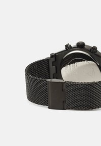 Reloj con correa de malla negra y caja de acero inoxidable. Presenta una esfera redonda, acentos plateados y un cierre grabado con el nombre de la marca.