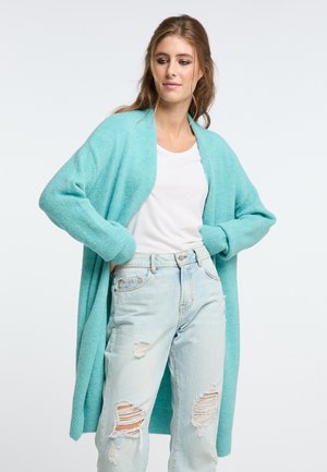 Turquoise cardigan van zacht, gestructureerd materiaal met een open voorkant, lange mouwen en geribde manchetten, gecombineerd met een wit T-shirt en versleten jeans.