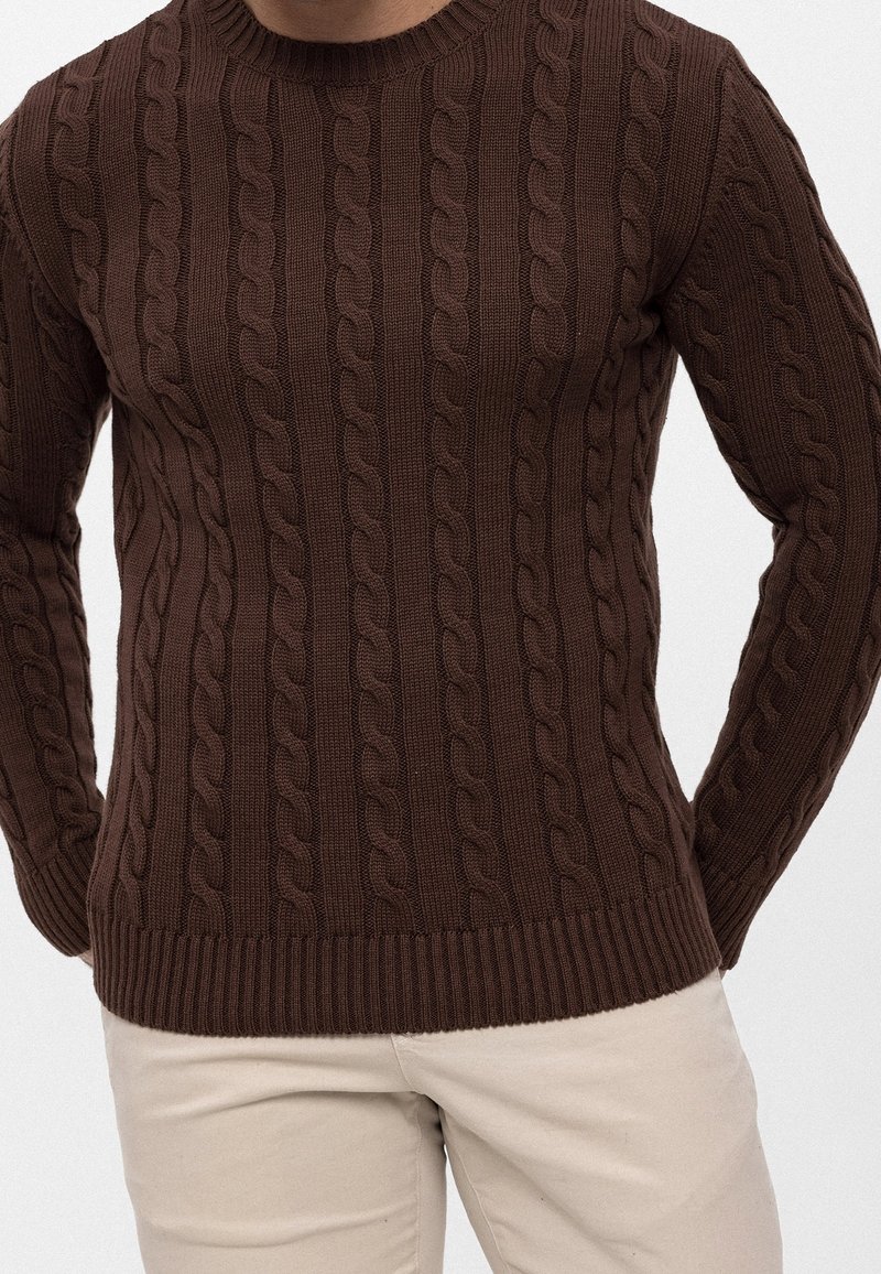Uomo che indossa un maglione a trecce marrone aderente con maniche lunghe, abbinato a pantaloni beige chiaro, su uno sfondo semplice.