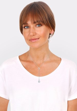 Silberne Halskette mit einem herzförmigen Anhänger und einem herabhängenden runden Charm, getragen über einem weißen T-Shirt. Glatte Textur, schlichtes Design.