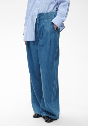 Personne portant un jean bleu taille haute à jambes larges avec plis, mains dans les poches, associé à une chemise à rayures bleu clair boutonnée et des chaussures noires.