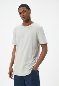 Camiseta de manga corta gris claro hecha de una tela texturizada con un cuello redondo. Combinada con unos vaqueros azul oscuro, manos en los bolsillos.