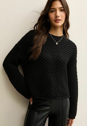 Schwarzer Strickpullover mit einem strukturierten Waffelmuster und langen Ärmeln, kombiniert mit enganliegenden schwarzen Hosen. Schlichter runder Ausschnitt und keine sichtbaren Verschlüsse.