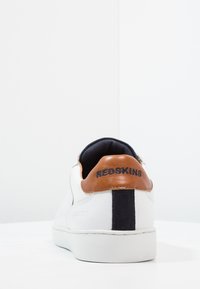 White sneakers with a textured leather upper, brown leather accent at the heel, and navy fabric lining. Features a rubber sole and branding.