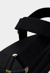 Μαύρο σανδάλι με φαρδιές υφασμάτινες λουρίδες και λεπτομέρειες από χρυσό μεταλλικό δαχτυλίδι, με κοντινή λήψη του κλεισίματος και της υφής του λουριού.