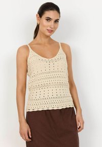 Top in cotone all'uncinetto di colore beige chiaro con orlo ondulato, caratterizzato da un motivo a maglia aperta e spalline sottili. Abbinato a una gonna marrone.