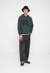 Young man wearing a dark green hoodie with graphic print, black bucket hat, wide black jeans, and black sneakers with white laces in plain studio.
