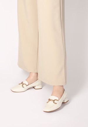 White leather block-heeled shoes with a square toe and gold chain detail across the instep, paired with beige wide-leg pants.