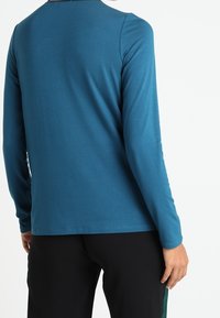 Pitkähihaisen puseron väri on teal, ja se on valmistettu sileästä kankaasta. Sitä koristaa pyöreä kaula-aukko sekä yksinkertainen ja siisti muotoilu ilman kuvioita tai koristeita.