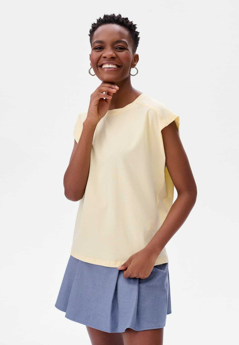 Lichtgele short-sleeve top van gladde stof, gecombineerd met een blauwe plooienrok. Ronde halslijn en subtiele stikdetails.