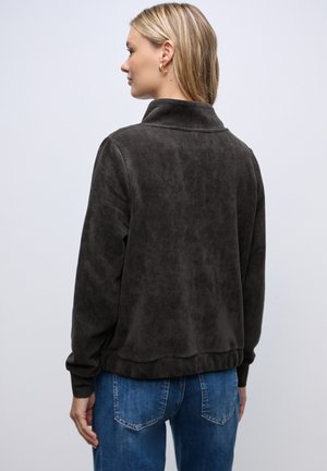Giacca in velluto a coste marrone scuro con colletto alto, polsini e orlo rigati. Indossata con jeans azzurri chiari, caratterizzati da una superficie testurizzata.
