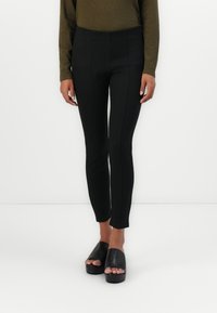 Persona che indossa pantaloni neri aderenti e scarpe con tacco aperto nere, in piedi contro uno sfondo bianco uniforme.