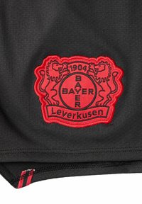 Schwarzer Sportstoff mit einem gestickten roten Wappen, das zwei Löwen zeigt, den Text "BAYER" und "Leverkusen" sowie einen dekorativen roten gestreiften Akzent.