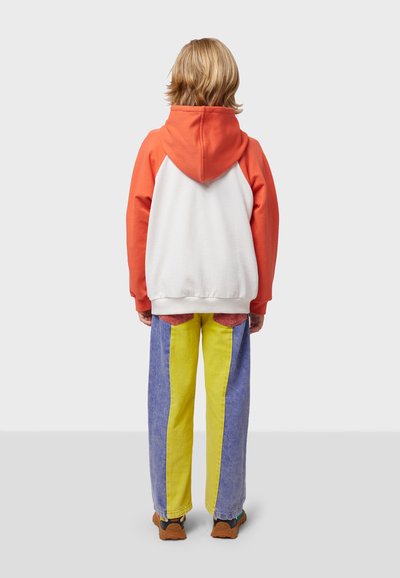 Sudadera colorida con mangas naranjas y cuerpo blanco, combinada con pantalones de mezclilla con rayas verticales moradas y amarillas. Zapatos marrones.