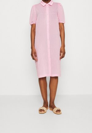 Persona con un vestido rosa hasta la rodilla abotonado con mangas cortas abullonadas y sandalias beige trenzadas sin cordones, de pie frente a un fondo blanco.