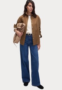 Veste en daim marron à boutons, pull blanc, jeans bleu large, tenant un sac texturé beige. Chaussures noires. Design simple et épuré.