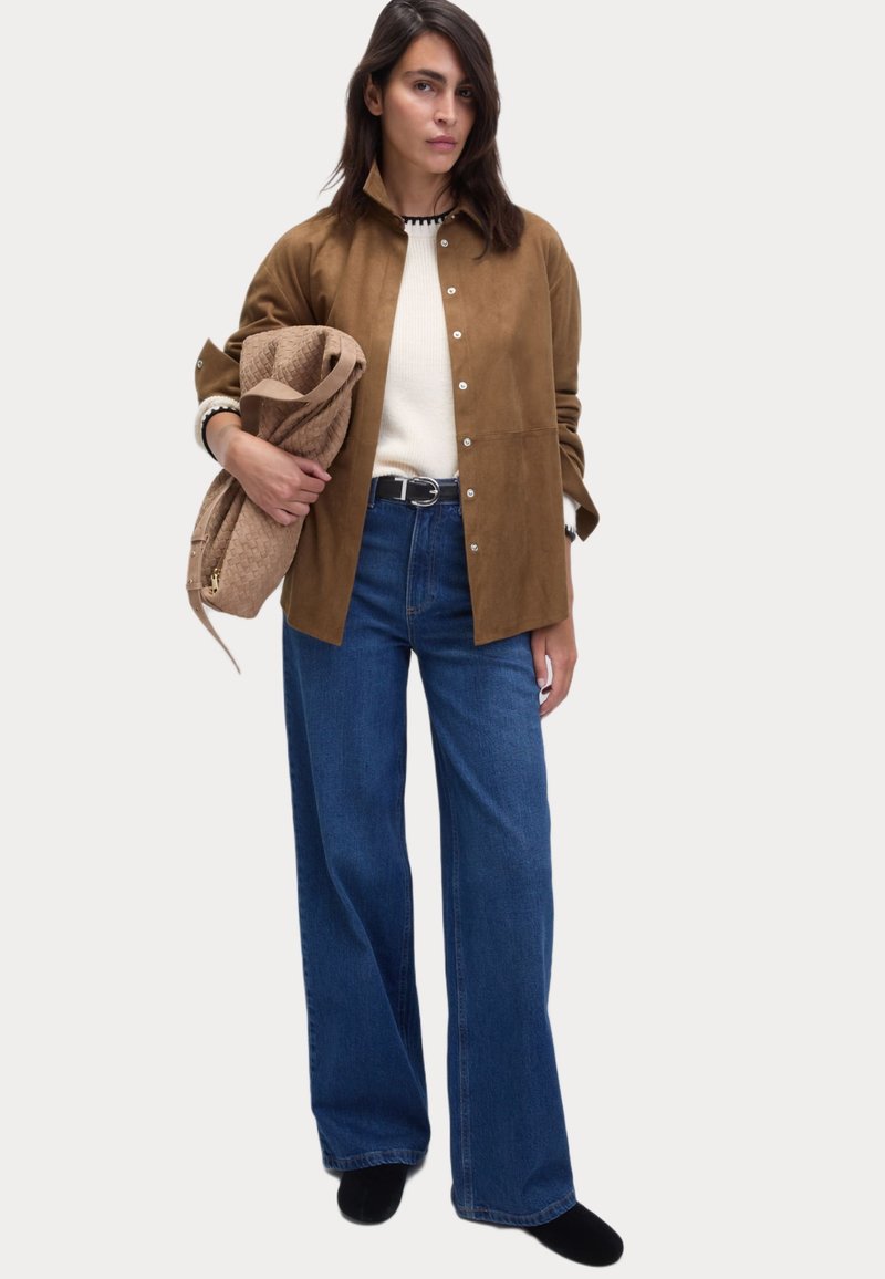 Veste en daim marron à boutons, pull blanc, jeans bleu large, tenant un sac texturé beige. Chaussures noires. Design simple et épuré.