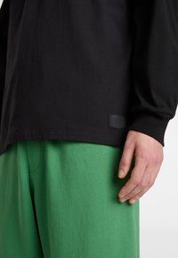 Schwarzes langärmliges T-Shirt aus Baumwolle, locker geschnitten mit einem strukturierten Logo-Patch; kombiniert mit leuchtend grünen Hosen.