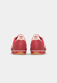 Zapatillas rojas con rayas blancas, hechas de ante y malla. Tacón redondeado, suela de goma y un logo de marca en la parte trasera.