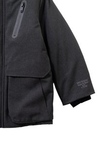 Donkergrijze winterjas van textuurstof, met grote zijzakken en ritssluitingdetails. De jas heeft branding op de mouw en een ontspannen pasvorm.