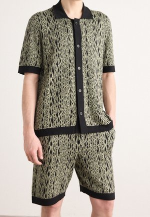 Man wearing matching green and black patterned short-sleeve button-up shirt and shorts with black trim on collar, cuffs, and hem.