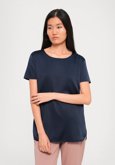 Woman with long black hair wearing a navy blue short-sleeve blouse and light pink pants, posing with one arm across her body.