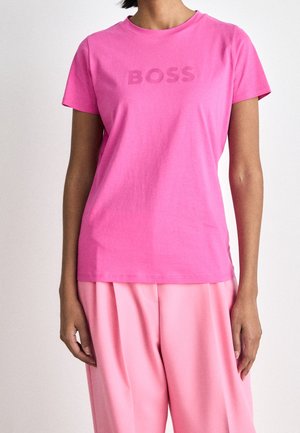 Osoba ubrana w jasnoróżową koszulkę z krótkim rękawem z napisem "BOSS" oraz luźne jasnoróżowe spodnie na tle jednolitym jasnym.