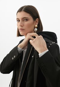 Musta pinstripe-puku, jossa on kultaiset ja hopeiset näyttävät korvakorut. Takissa on vetoketjullinen etuosa ja helmetyksityiskohtia olkapäillä. Alle on lisätty hupullinen kerros.