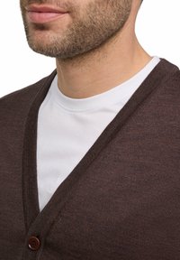 Brauner Cardigan mit V-Ausschnitt und einem Knopfverschluss, schichtweise über einem weißen Rundhalsausschnitt-Shirt getragen. Texturiertes Strickmaterial sichtbar.