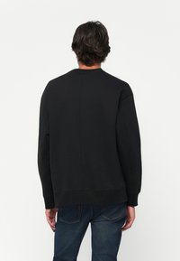 Άντρας που φοράει μαύρη μπλούζα με μακριά μανίκια και μπλε τζιν, απεικονιζόμενος από πίσω σε απλή λευκή φόντο.