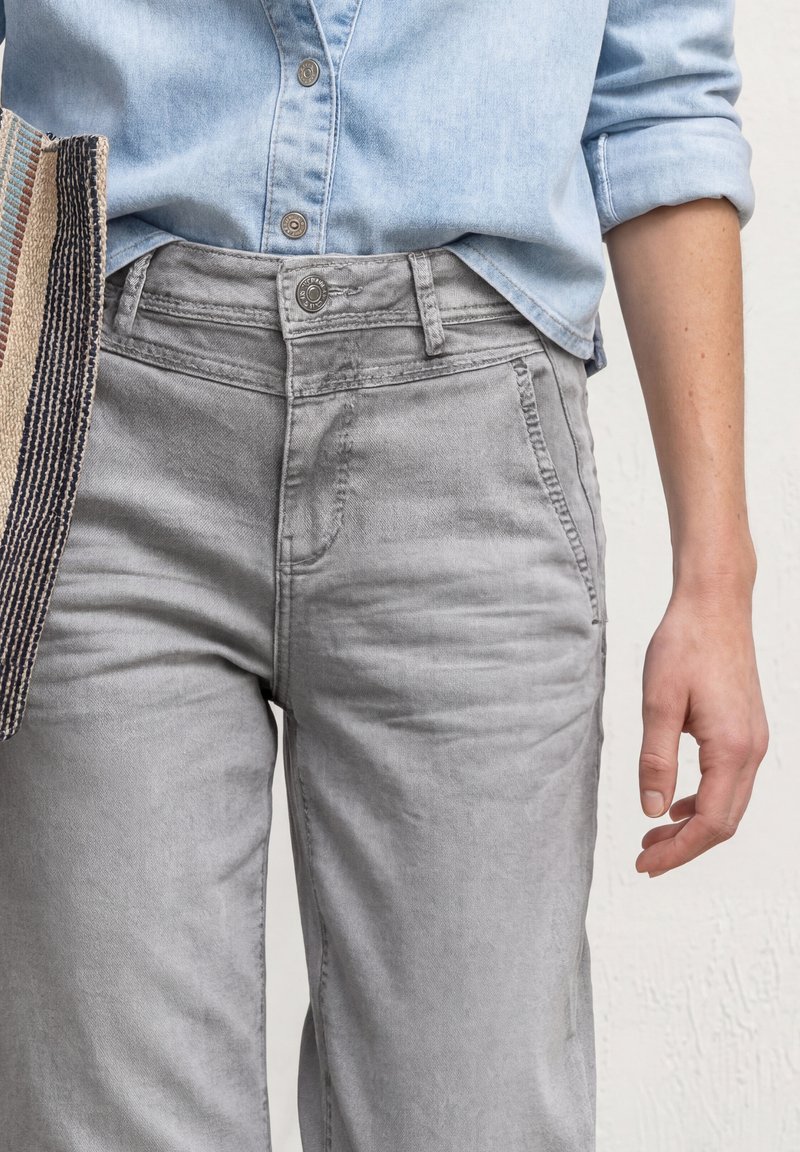 Personne portant une chemise bleu clair boutonnée rentrée dans un jean gris taille haute avec un sac fourre-tout rayé sur le côté gauche.