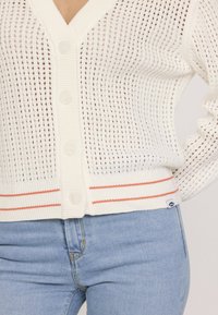 Cardigan léger en tricot blanc avec des côtes verticales, cinq boutons et une accent rayé orange à l'ourlet. Porté sur un jean bleu.