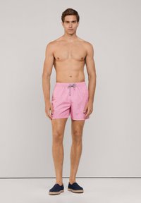 Różowe kąpielówki w wzory z elastycznym pasem i sznurkiem. Model stoi boso, w ciemnych butach wsuwanych.