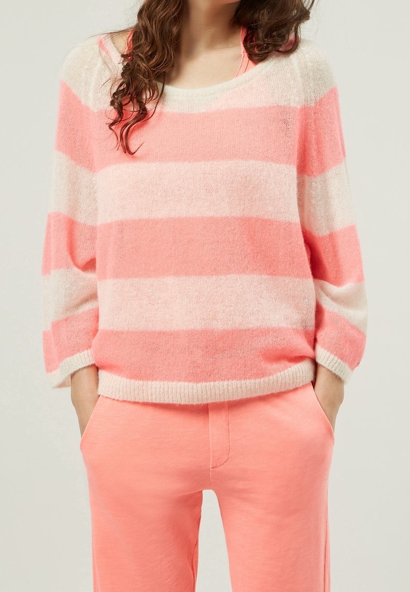 Femme portant un pull rayé rose et blanc avec des manches trois-quarts amples et un pantalon rose, mains dans les poches.