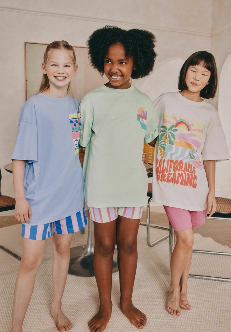 Tre børn har store t-shirts på i pastelnuancer: blå, grøn og hvid med farverige tryk, samt stribede shorts.