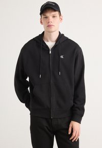 Sudadera con cremallera negra hecha de un tejido suave. Presenta un bolsillo delantero, capucha con cordones y un logo bordado en el lado izquierdo del pecho.